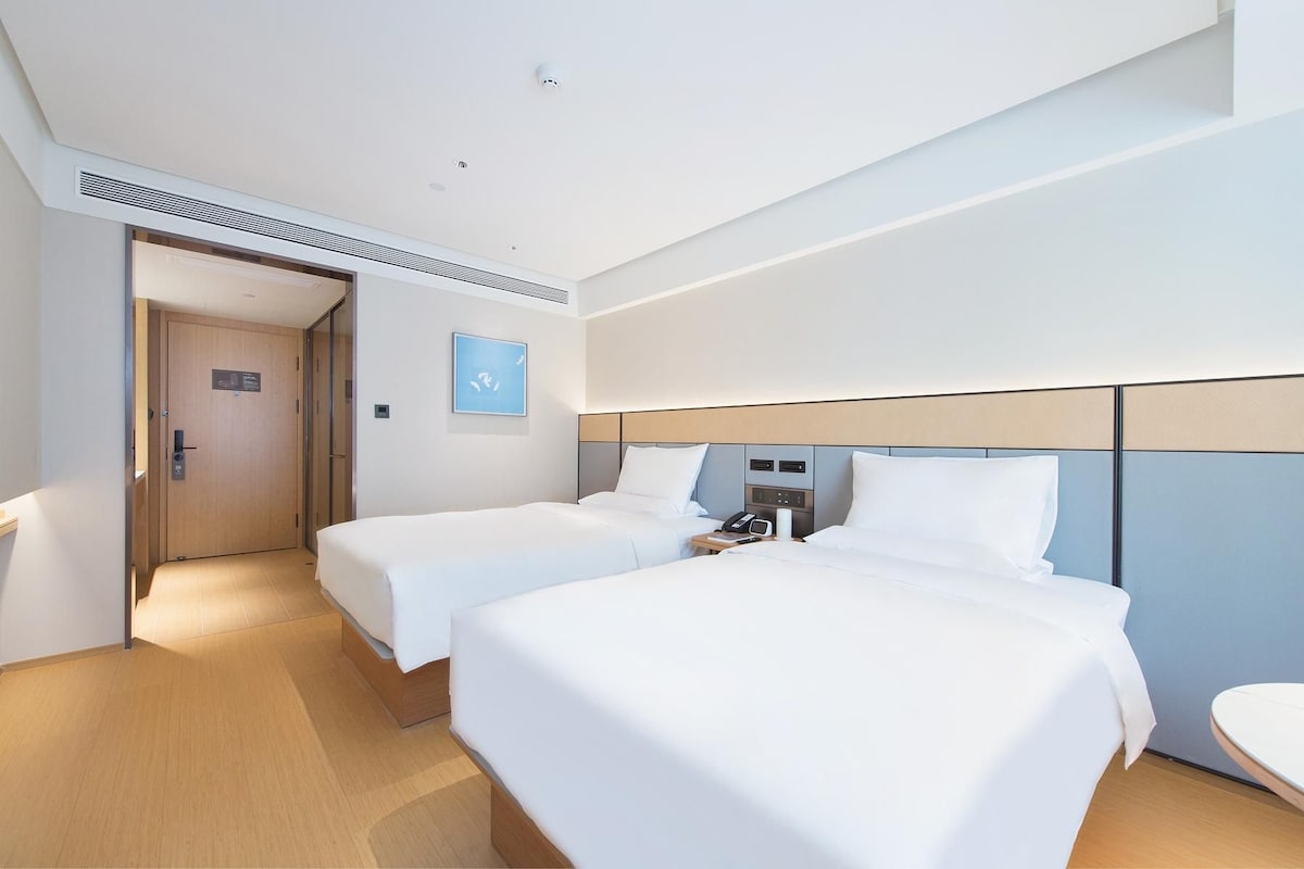 Quarto de hotel com duas camas, uma mesa de cabeceira com telefone e abajur, uma porta com placa e uma pequena obra de arte na parede.