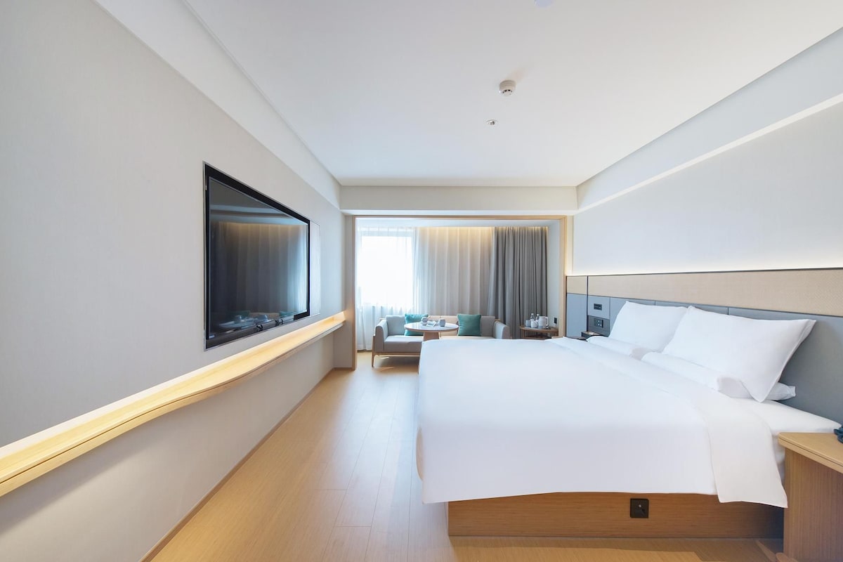 Quarto de hotel com cama, mesa de cabeceira, televisão e vista para o corredor.