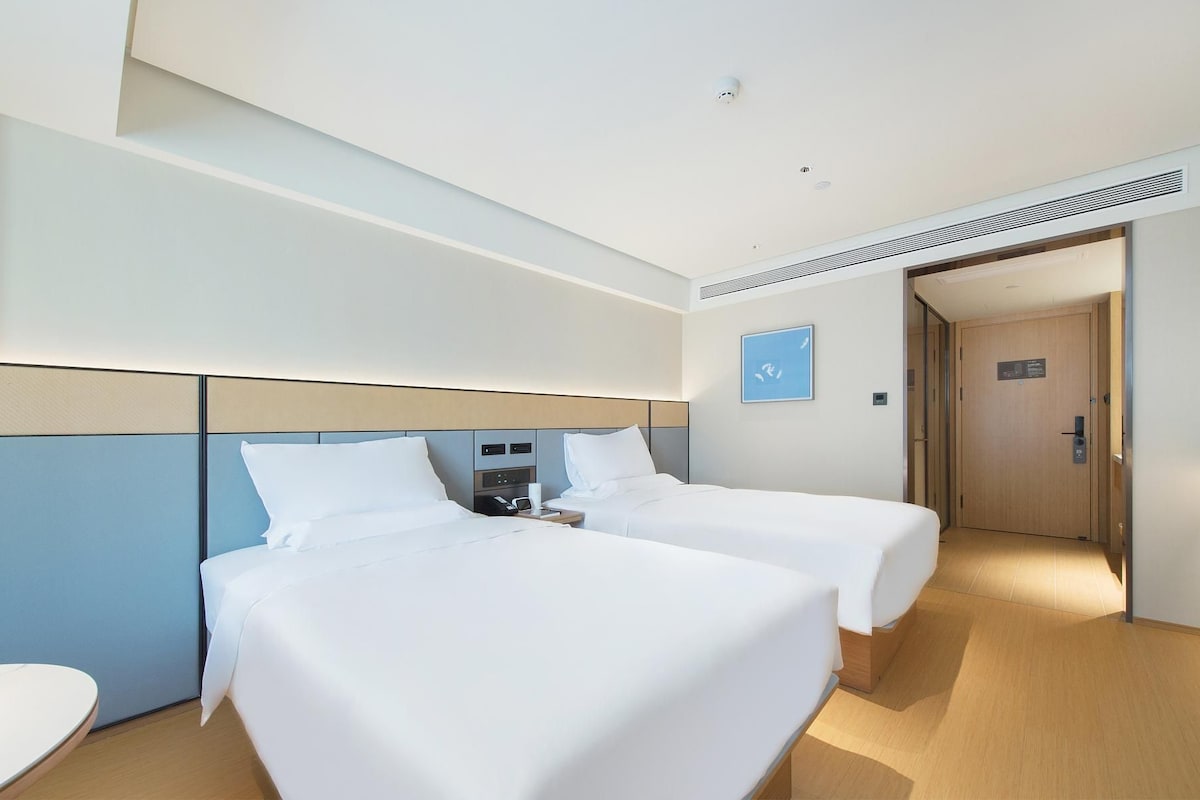 Quarto de hotel com duas camas, uma mesinha de cabeceira, um telefone fixado na parede e uma porta que dá para outro cômodo.