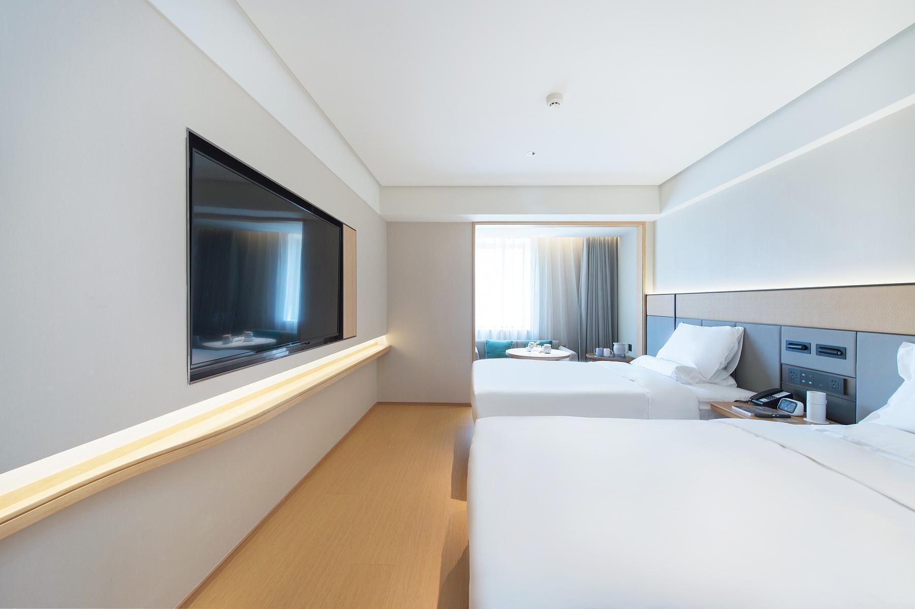 A hotel room with a large TV, a bed, a bedside table with a phone, and a window with curtains.