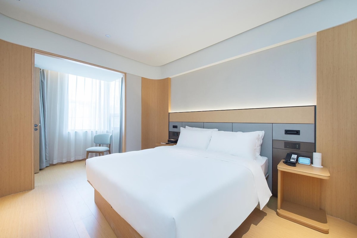 Quarto de hotel moderno com uma cama grande, mesas de cabeceira, uma cadeira e vista para o exterior.