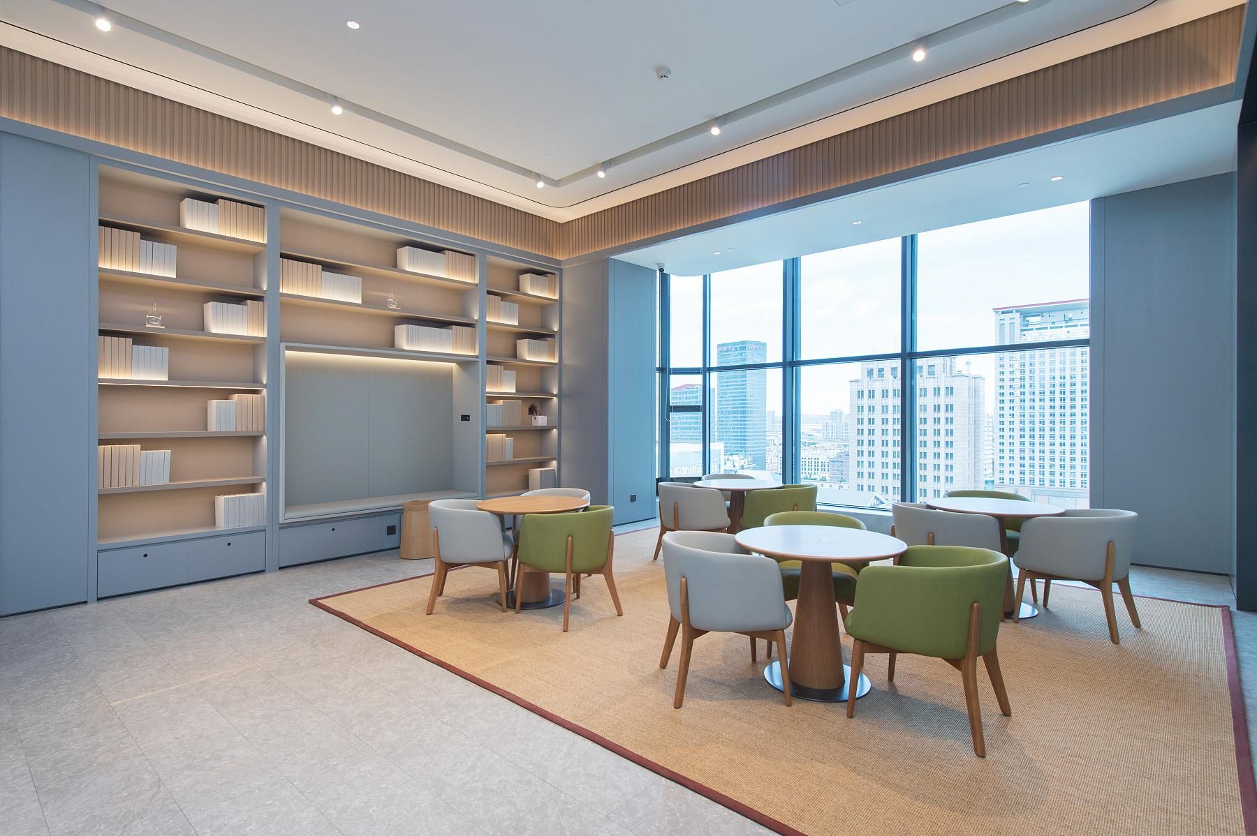 A modern library with bookshelves, round tables, and chairs, large windows with a city view.