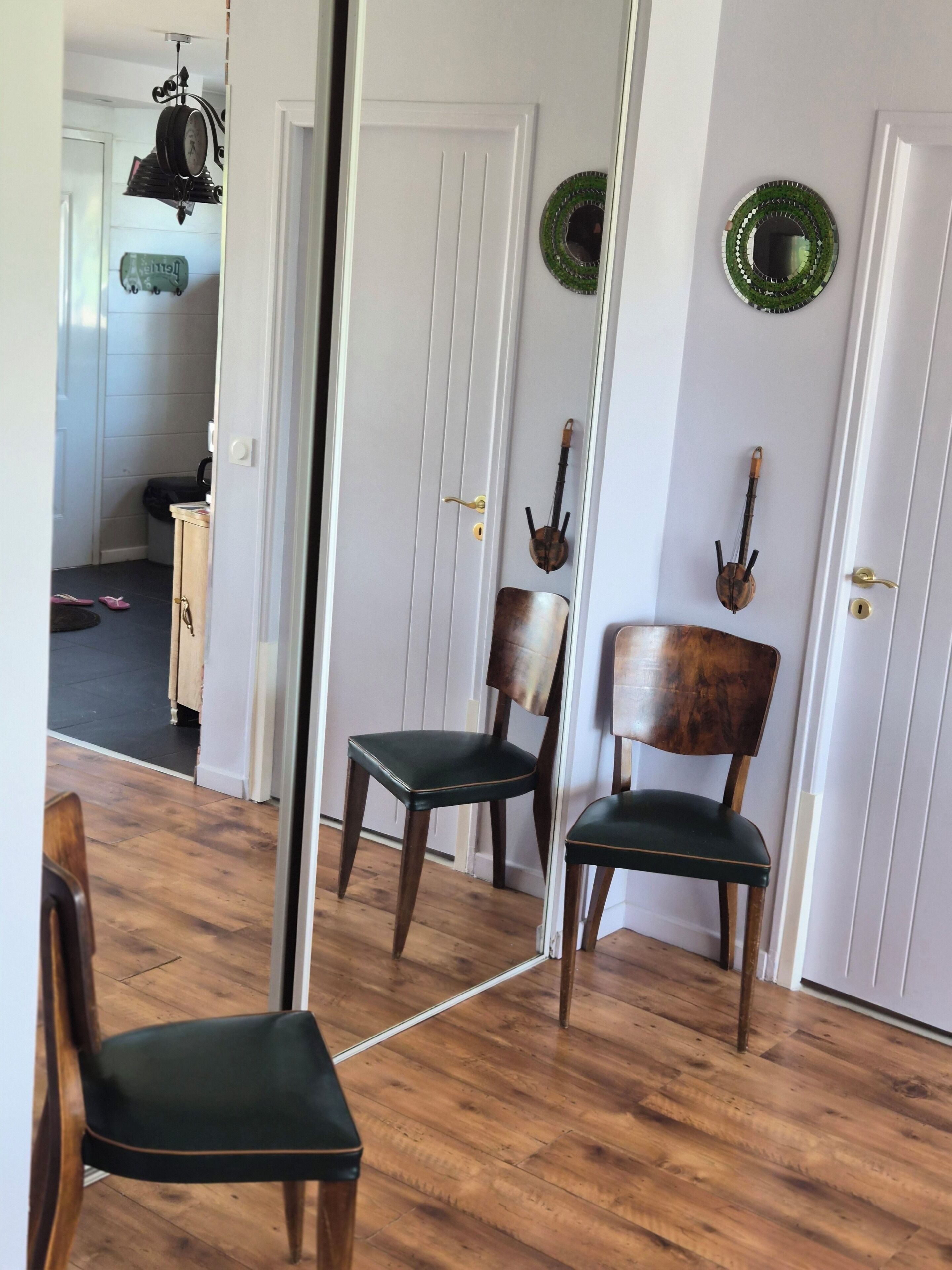 Une pièce avec un plancher en bois, un fauteuil recouvert d’un tissu vert, une chaise en bois, une porte blanche et un miroir qui reflète la pièce.