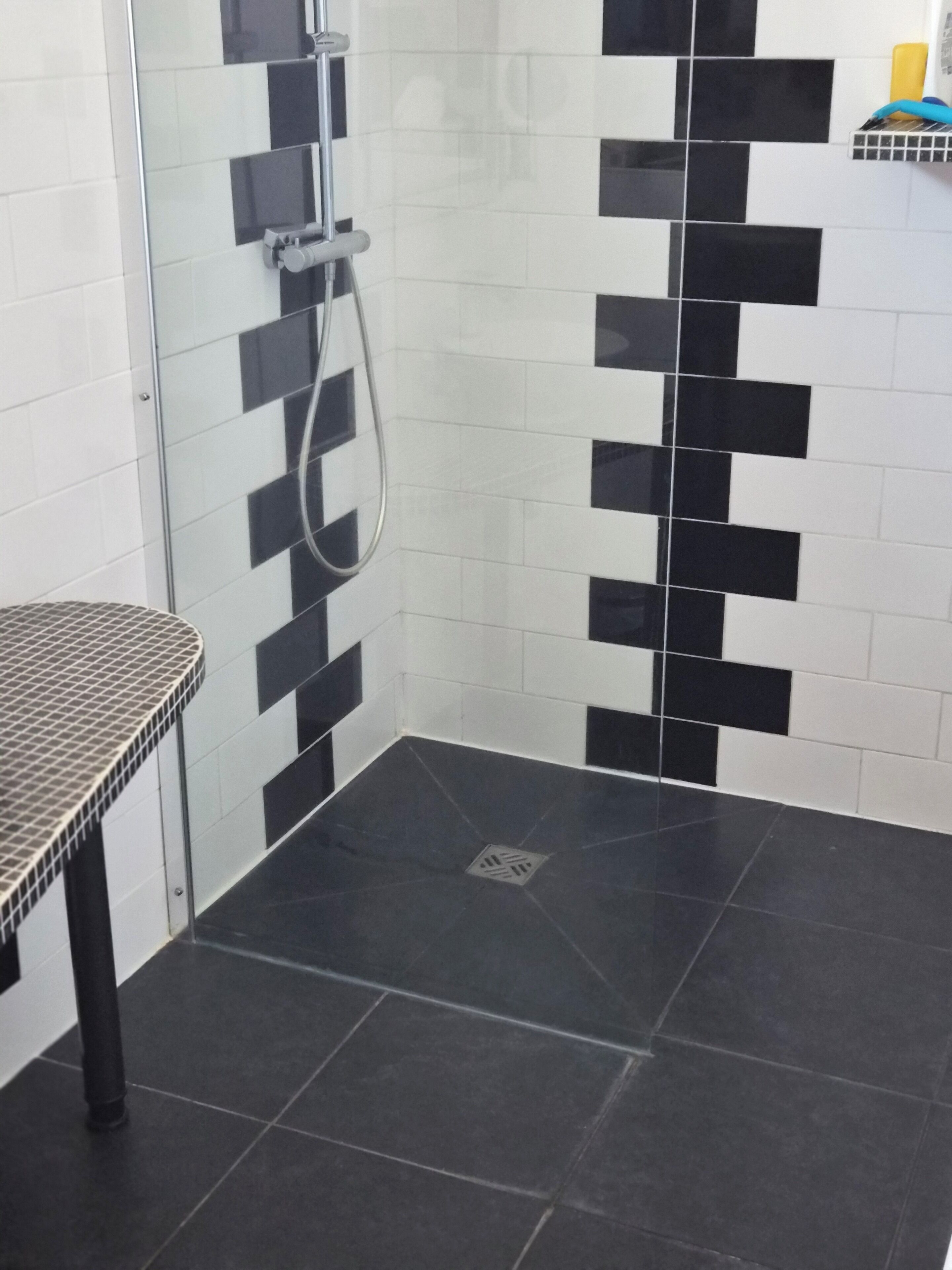 Une salle de bain avec des carreaux noirs et blancs disposés en motif à damier, une douche avec une porte en verre et une petite table dont le plateau est constitué d’une grille métallique.