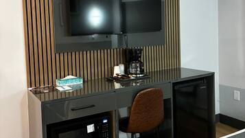 A modern hotel room with a flat-screen TV mounted on a wooden panel wall, a compact kitchenette with a microwave, and a small refrigerator.