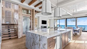 Une cuisine moderne dotée d’un grand îlot en marbre, de poutres en bois et offrant une vue sur la mer.