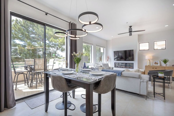 Uma sala de jantar moderna com mesa e cadeiras de vidro, ventoinha no teto e vista para o exterior.