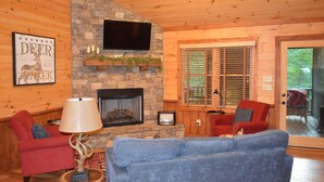 A cozy cabin interior with a stone fireplace, a television mounted above it, and a comfortable seating area with a blue sofa and red armchairs. - Cozy Cabin for 4 with 2 King beds, 2 full baths, 2 fireplaces and upscale Decor! (Morganton)