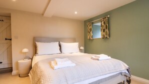 Una cama bien hecha con sábanas blancas y una colcha acolchada, una lámpara de noche y una ventana con cortinas estampadas con hojas.