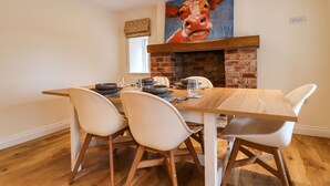Un comedor con chimenea de ladrillo, un cuadro grande de una vaca y una mesa de madera con sillas blancas.