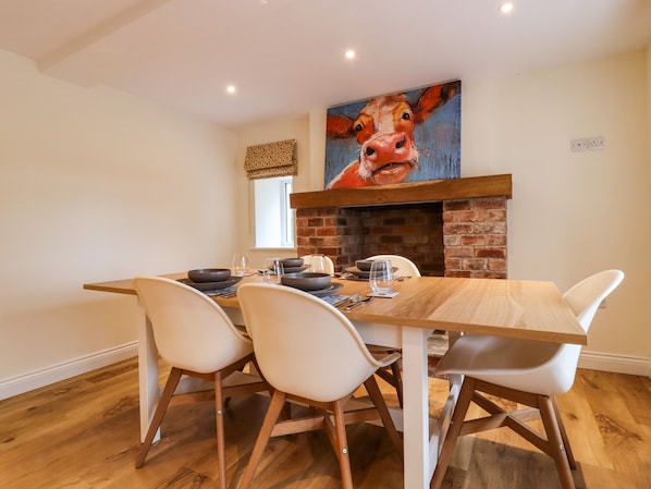A dining area with a brick fireplace, a large painting of a cow, and a wooden table with white chairs.