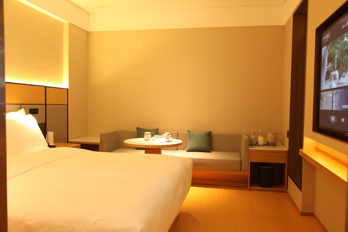 A hotel room with a bed, a sofa, a small table with tea set, a TV on the wall, and a trash bin.