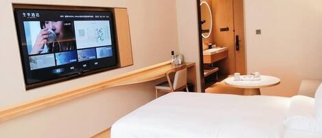 Habitación de hotel con televisor montado en la pared mostrando un menú, una cama, un escritorio con una silla, una pequeña mesa redonda con una taza y un platillo, y un espejo sobre la puerta.