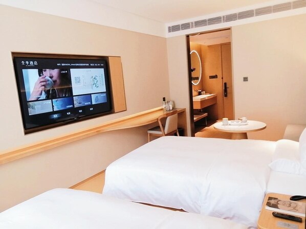 Ein Hotelzimmer mit einem an der Wand befestigten Fernseher, der eine Speisekarte zeigt, einem Bett, einem Schreibtisch mit Stuhl, einem kleinen runden Tisch mit Tasse und Untertasse sowie einem Spiegel über der Tür.