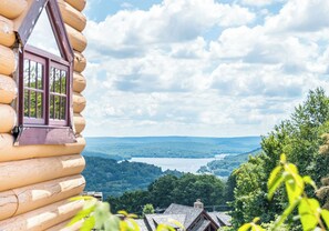 A log cabin with a window overlooking a lake and forested hills.