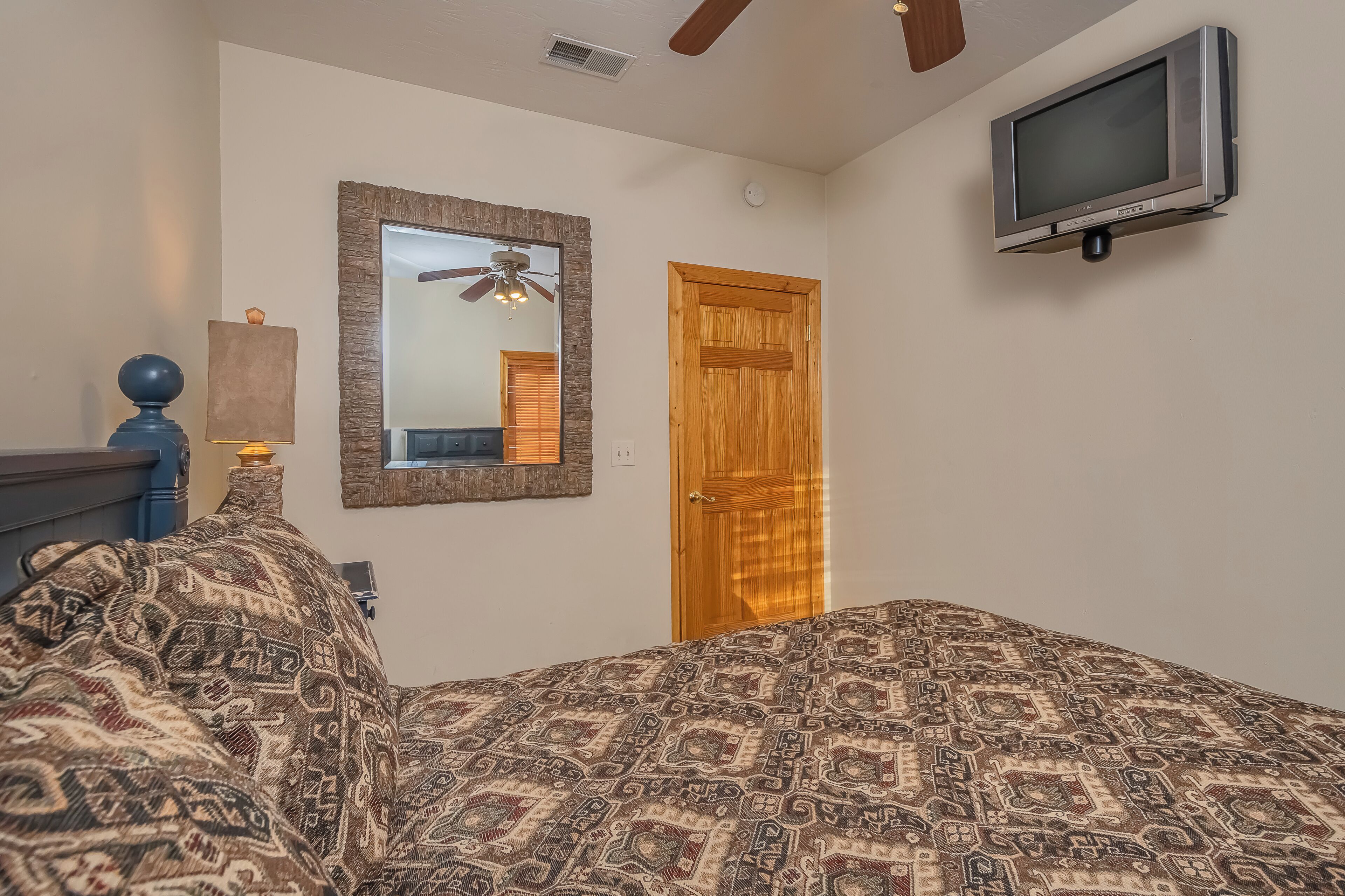 Ein Schlafzimmer mit gestreiftem Bettbezug, Deckenventilator, angebrachtem Fernseher und einer Holztür.
