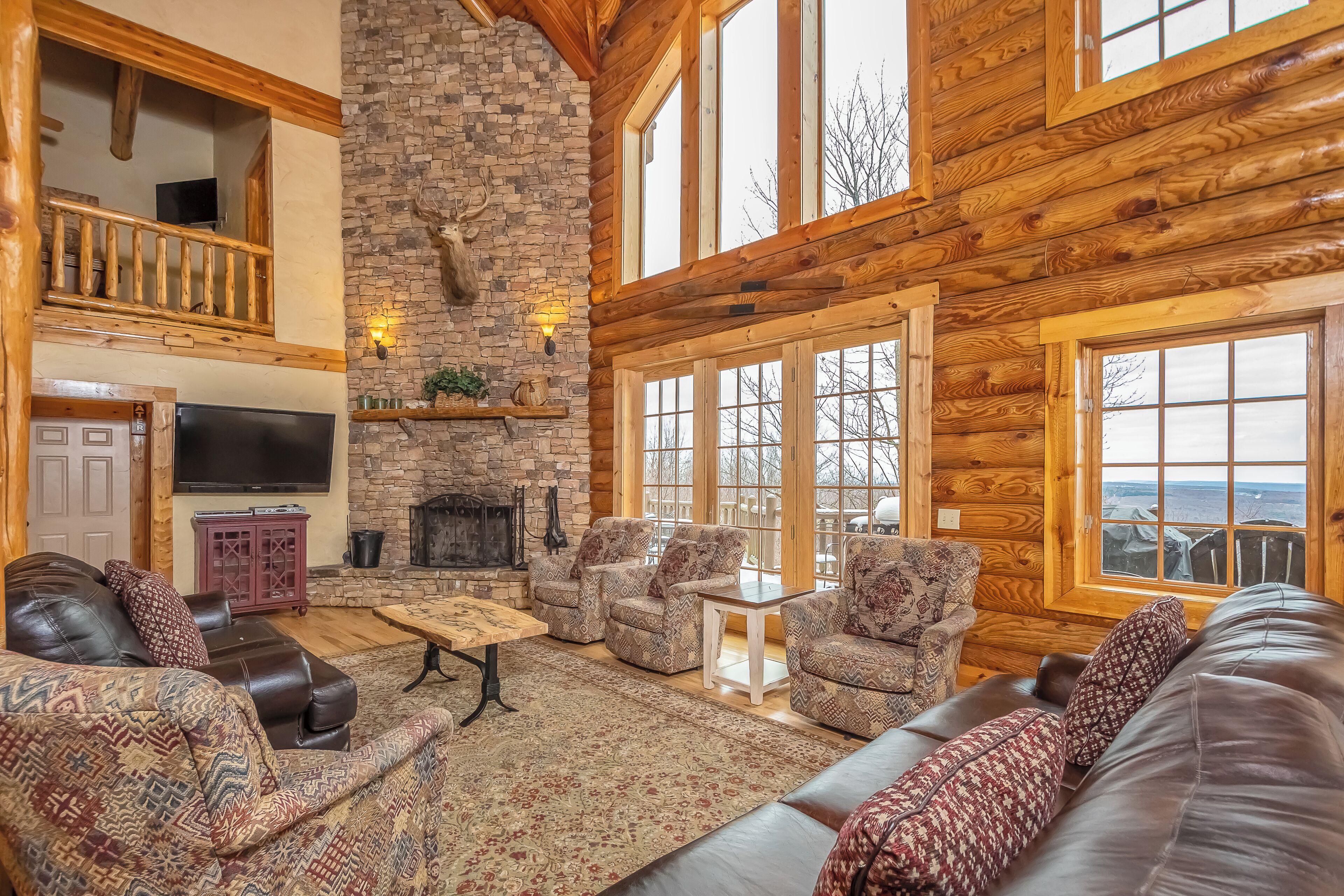 L’intérieur d’une cabane en rondins, avec une cheminée en pierre, des canapés en cuir, des fauteuils à motifs et de grandes fenêtres offrant une vue panoramique.