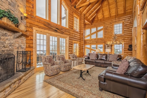 A log cabin interior with a stone fireplace, large windows, leather sofas, and a chandelier.