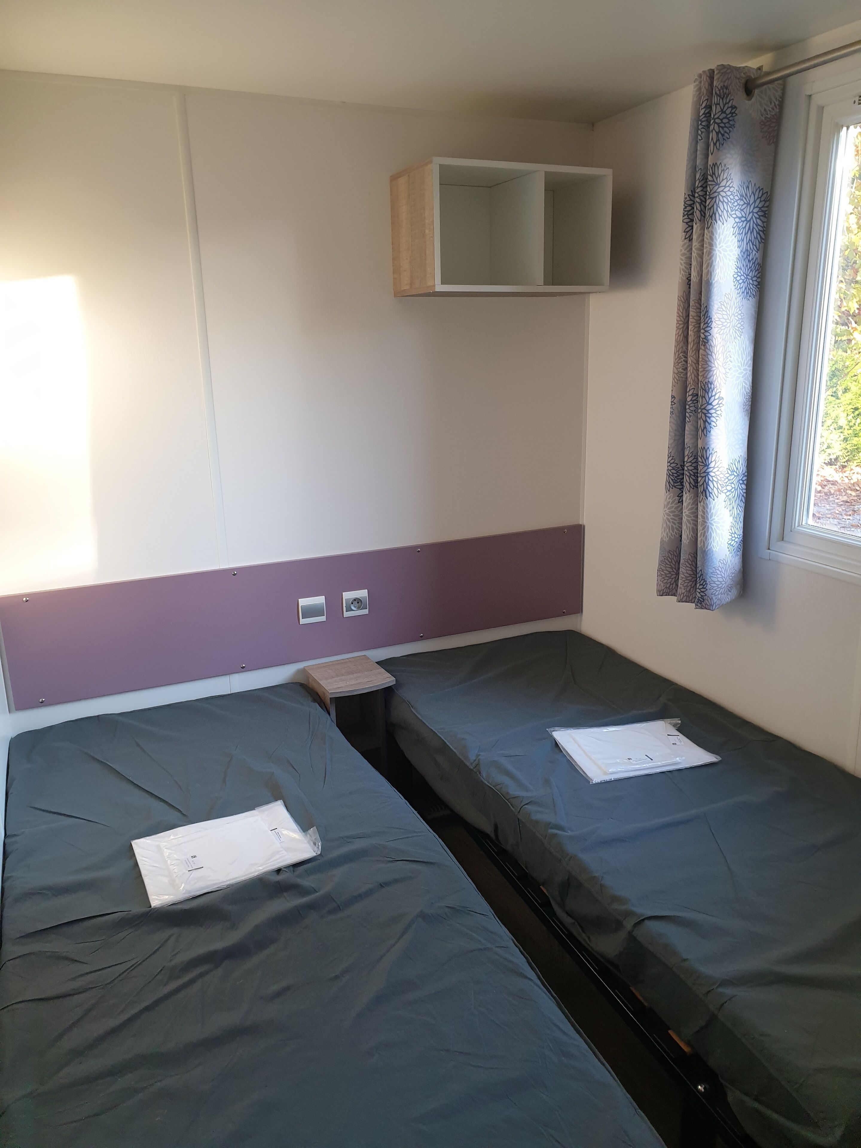 Ein Zimmer mit zwei Betten, einem kleinen Tisch, einem Regal und einem Fenster mit Vorhängen.