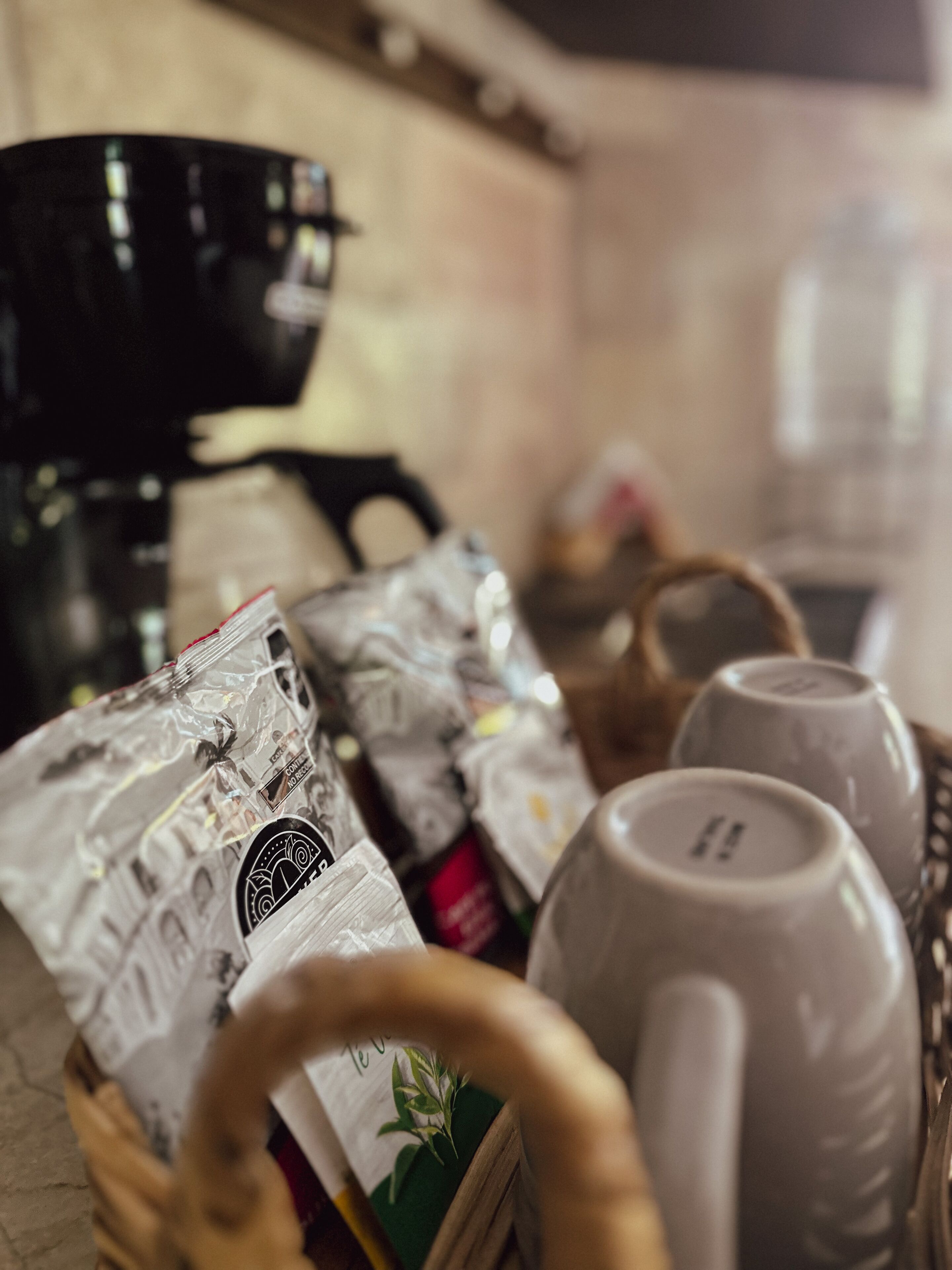 Un área de cocina con cafetera, un cesto de cápsulas de café y un juego de tazas blancas.