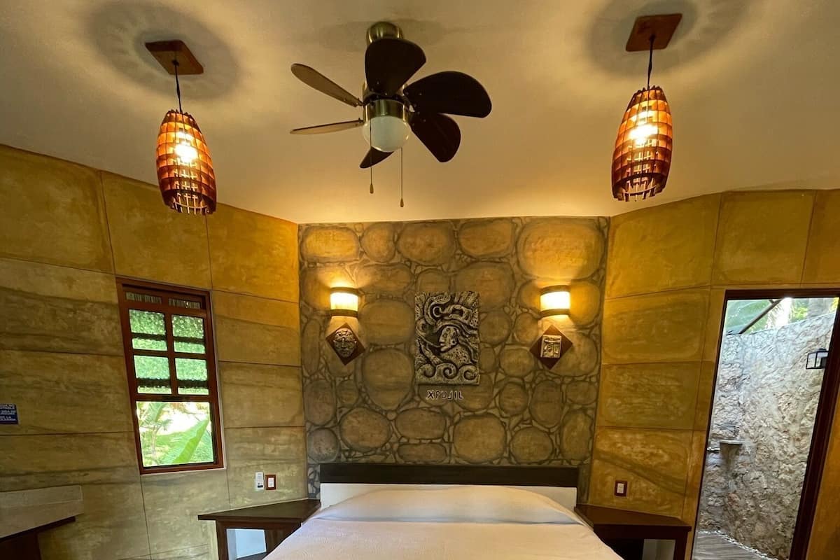 Un dormitorio con cama, ventilador de techo, dos luces fijadas a la pared, pared de piedra, ventana y puerta.