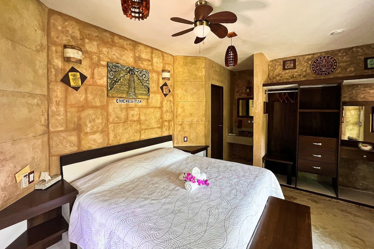 Una habitación de hotel con cama, ventilador de techo, una obra de arte colgada en la pared, espejo y armario.