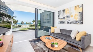 A modern living room with a grey sofa, wooden coffee table, and a glass door leading to a balcony with a view of the ocean and palm trees. - Condo Aria Ocean 306 (Nuevo Nayarit)