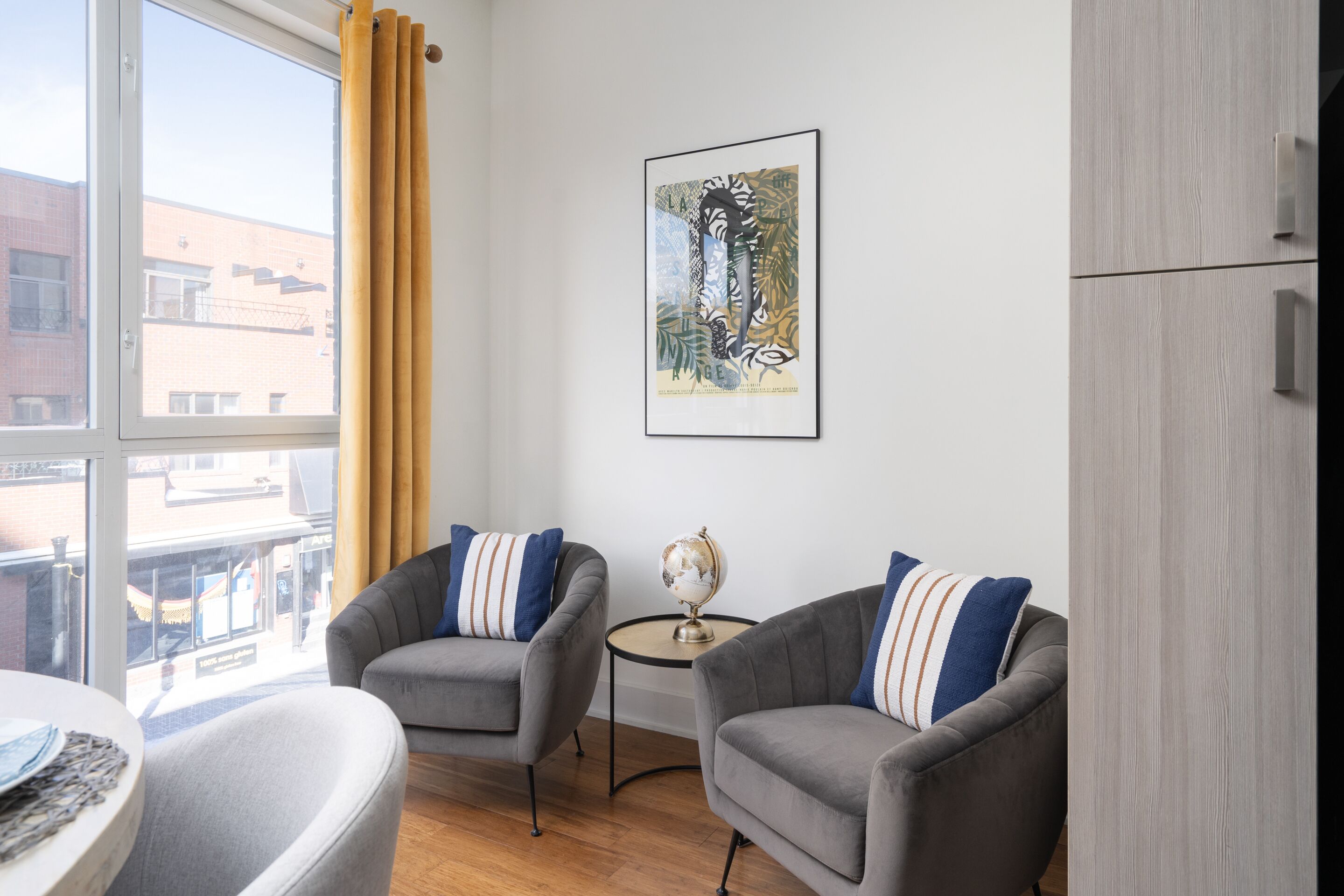 A modern living room with two grey armchairs, a small round table with a globe, and a framed artwork on the wall.