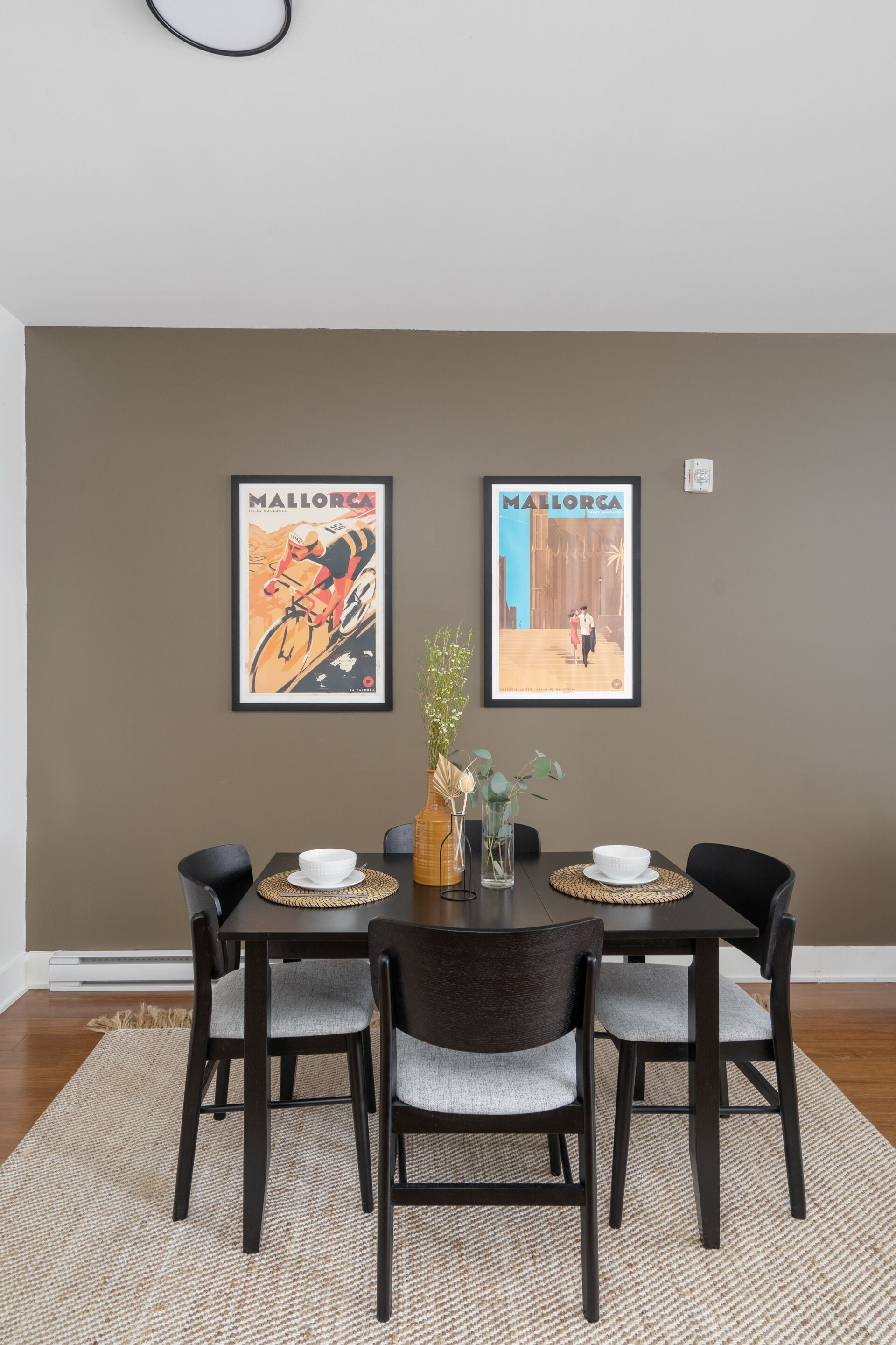 A dining area with a dark brown wall, two framed posters, a round table with four chairs, a vase with flowers, and a woven placemat.