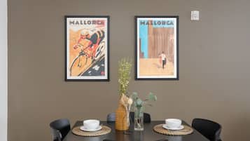 A dining area with a dark brown wall, two framed posters, a round table with four chairs, a vase with flowers, and a woven placemat.