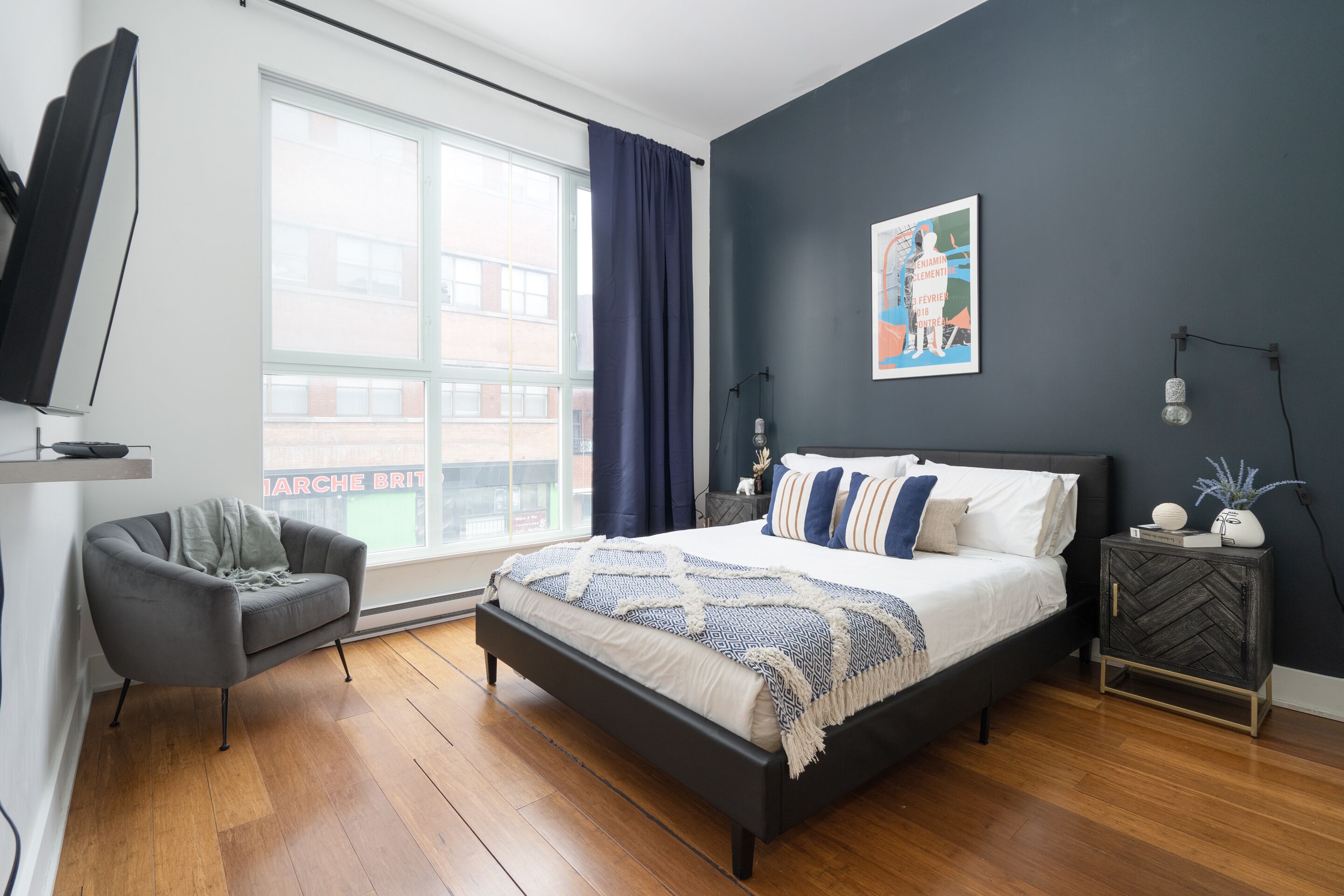 Une chambre moderne avec un grand lit, un fauteuil gris, une table de chevet avec une lampe et une fenêtre avec des rideaux.
