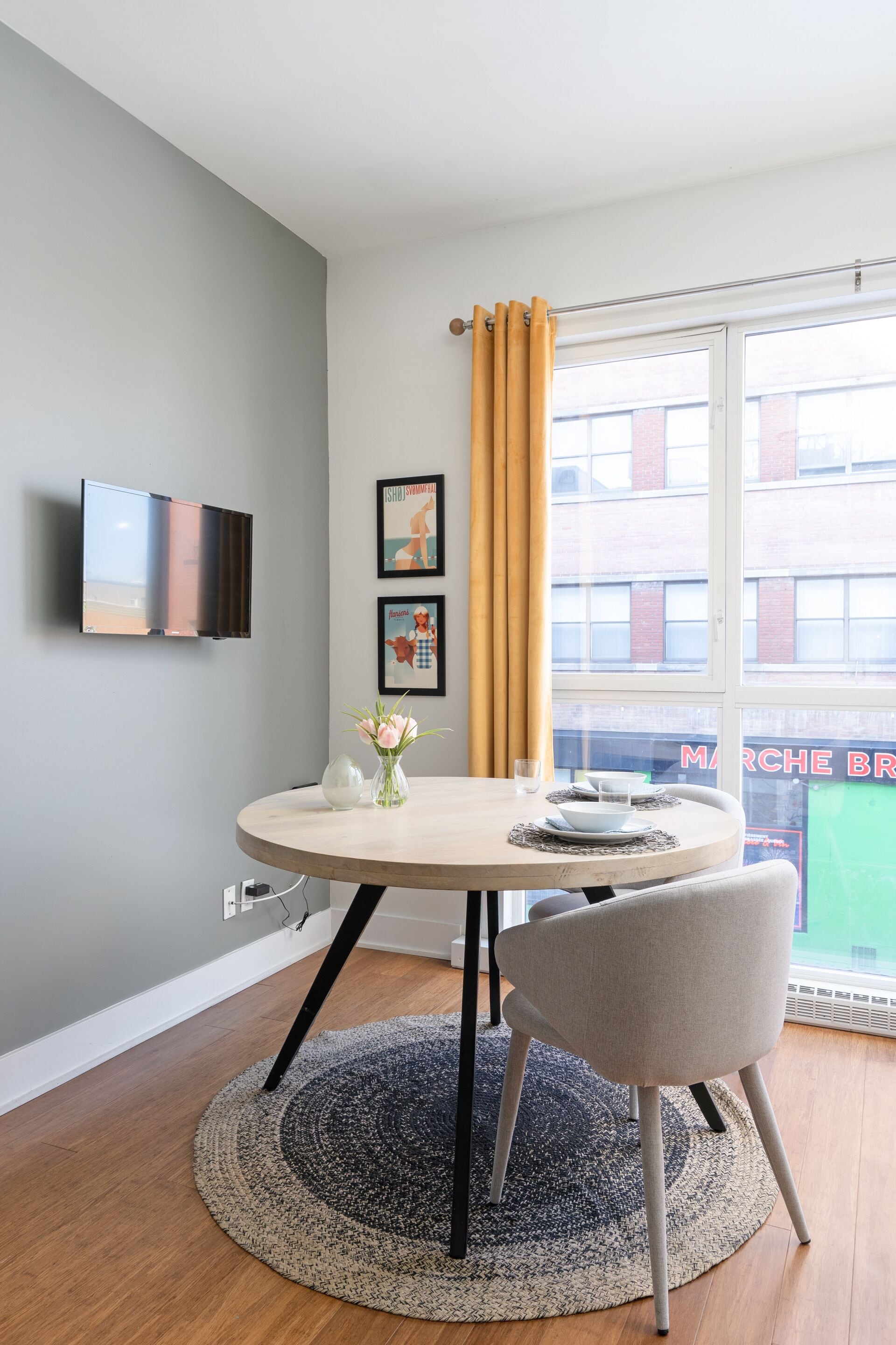 A modern dining area with a round table, a single chair, a vase with flowers, a flat-screen TV mounted on the wall, and a window with yellow curtains.