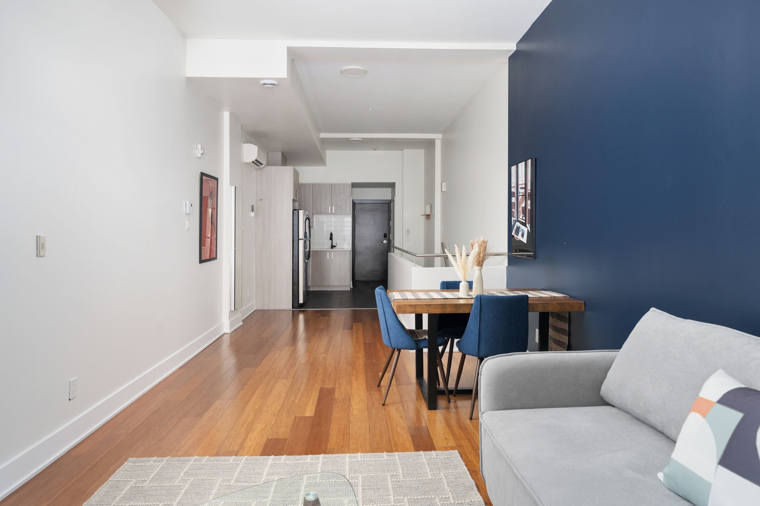 Una sala de estar moderna con un sofá gris, una mesa de comedor con sillas azules y una cocina al fondo.
