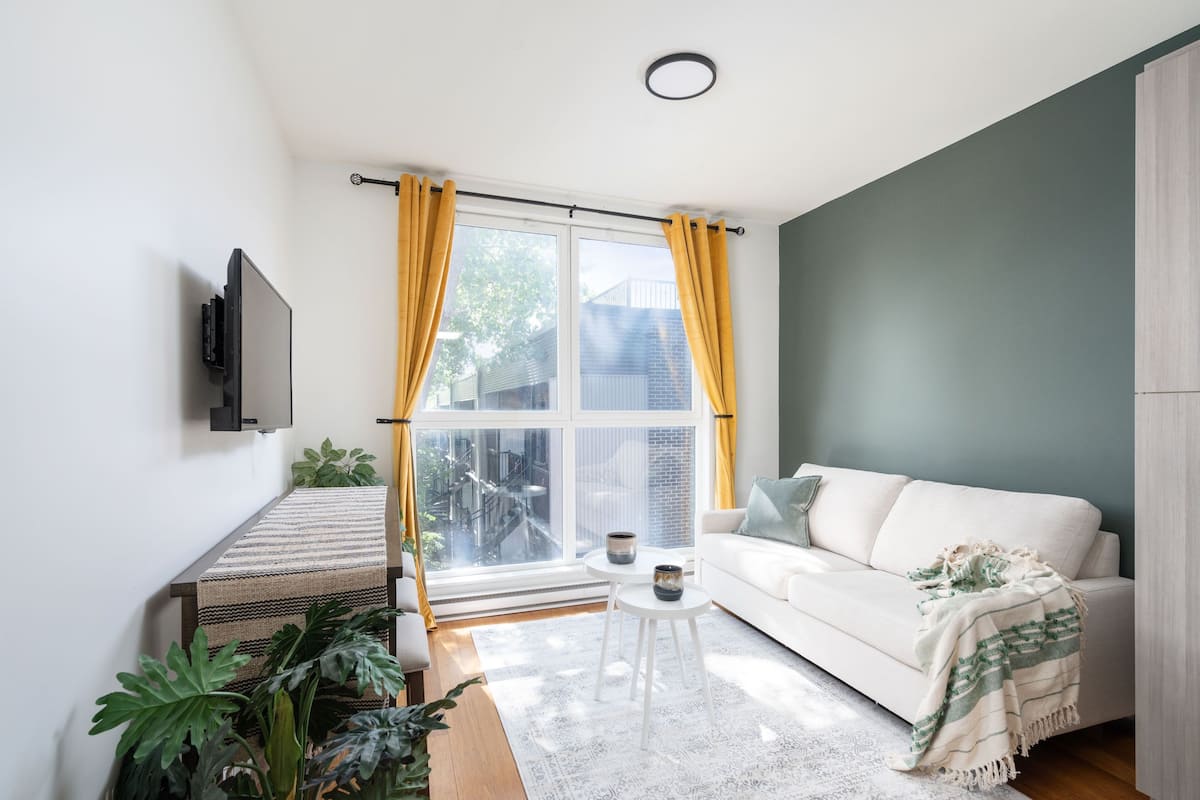 Una sala de estar moderna con un sofá, una mesa de centro, un televisor montado en la pared y un gran ventanal con cortinas.