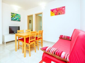 Una habitación con una mesa de comedor de madera, cuatro sillas, un sofá rojo y un televisor sobre un soporte blanco.