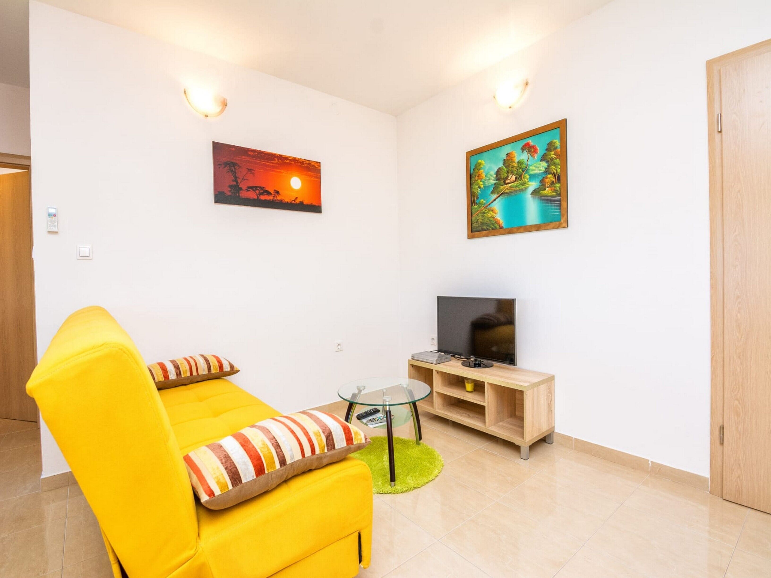 A bright yellow sofa with striped pillows, a flat-screen TV on a wooden stand, and two wall-mounted paintings depicting a sunset and a landscape.