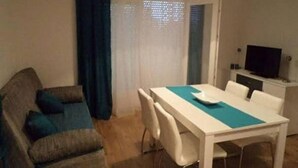 Ein kleiner, gut beleuchteter Raum mit Esstisch, Stühlen, einer Couch, einem Fernseher und einem Fenster mit Vorhängen.