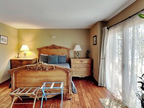 Une chambre à coucher avec un lit en bois, deux tables de chevet avec des lampes, une commode et une fenêtre ornée de rideaux légers.