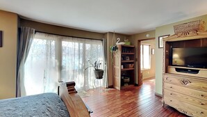 A spacious room with a large bed, wooden flooring, a television on a wooden cabinet, and a window with sheer curtains. - Blue Heron by Taylor Made Deep Creek (McHenry)