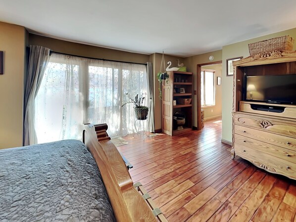 Une pièce spacieuse avec un grand lit, un plancher en bois, un téléviseur posé sur un meuble en bois et une fenêtre ornée de rideaux légers.