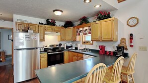 A kitchen with wooden cabinets, a stainless steel refrigerator, a microwave, a stove, and a coffee maker. There are wooden chairs around a central island. - Black Bear Retreat by Taylor Made Deep Creek (McHenry)