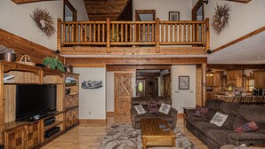 A spacious living room with a large TV, wooden furniture, a balcony, and a kitchen area.