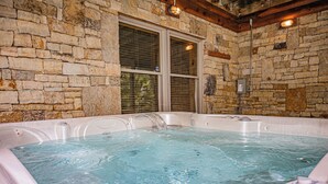 A stone-walled room with a large bathtub filled with water, a wooden ceiling, and a window with blinds.
