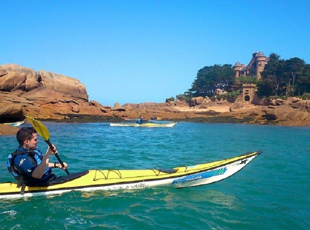 A kayaker in a yellow kayak on calm waters with a rocky shoreline and a castle-like structure in the background.