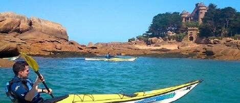 A kayaker in a yellow kayak on calm waters with a rocky shoreline and a castle-like structure in the background.
