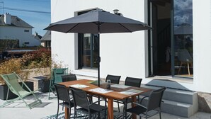 Un patio con una mesa y sillas de madera, un paraguas grande y vistas a un edificio blanco con varias ventanas.