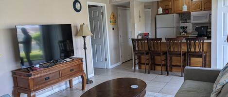 A living room with a TV, wooden furniture, a sofa, and a kitchen area with a refrigerator and microwave.
