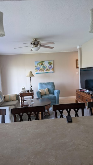 Uma sala de estar com ventilador de teto, um quadro com casas, sofá, mesa e televisão.