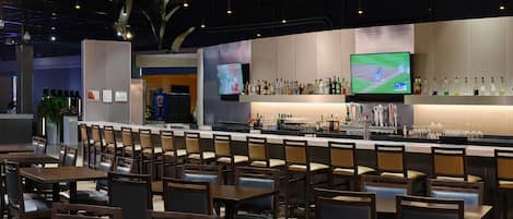 A bar area with multiple seating arrangements, a television screen, and a variety of bottles on the shelves.