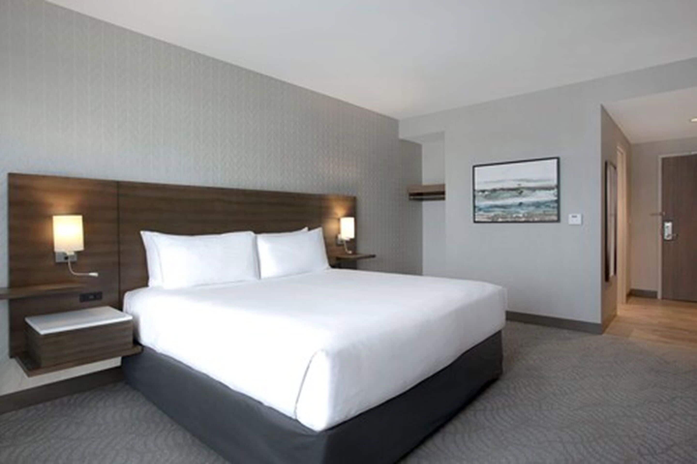 A modern hotel room with a large bed, two bedside tables with lamps, a framed picture on the wall, and a wooden door.